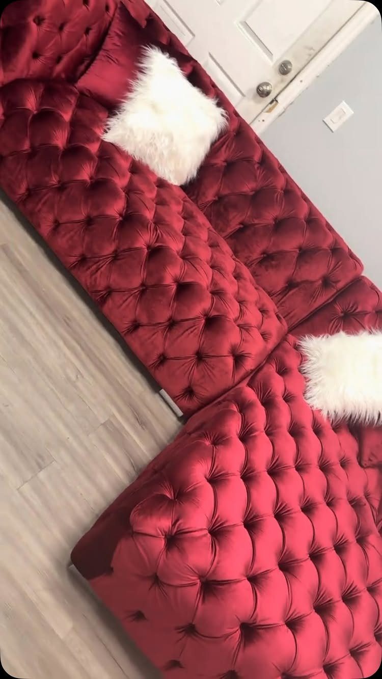 Red velvet tufted sectional sofa with white fluffy pillows on light wood flooring.