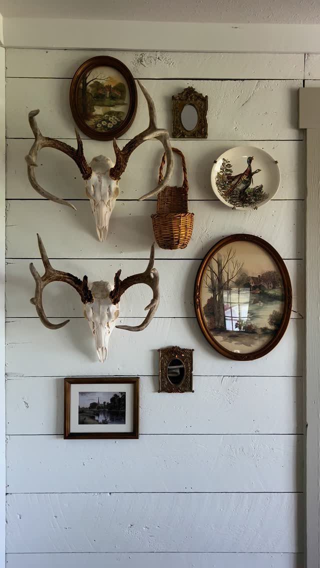 Gallery wall with deer antlers, landscape paintings, and mirrors on white wooden planks.