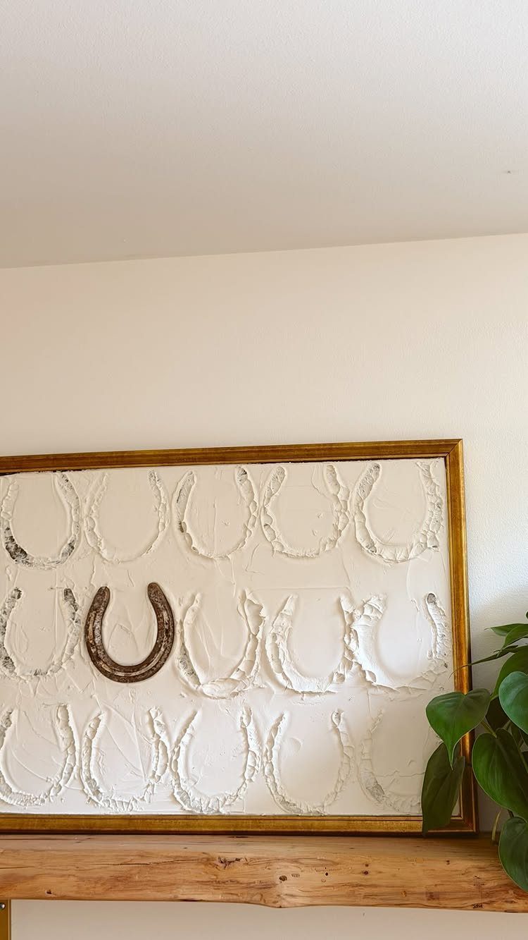 Framed horseshoe art with white texture and one metal horseshoe on wood shelf.