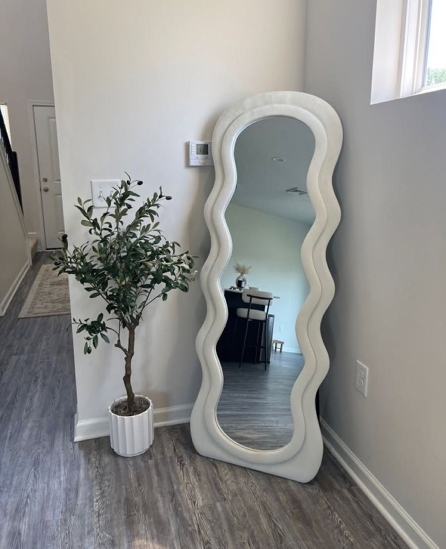 Wavy white full-length mirror leaning against a grey wall next to a potted plant
