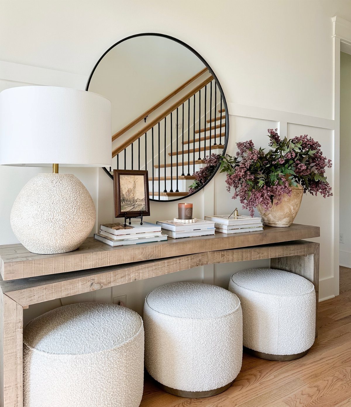 Light wood console table with white poufs, a round mirror, and floral decor.