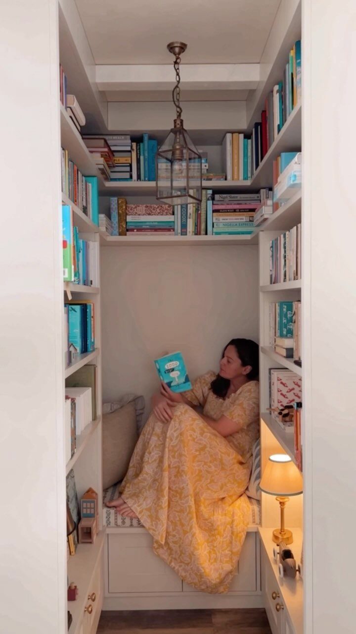 Woman reading in a white built-in reading nook surrounded by bookshelves and lighting.