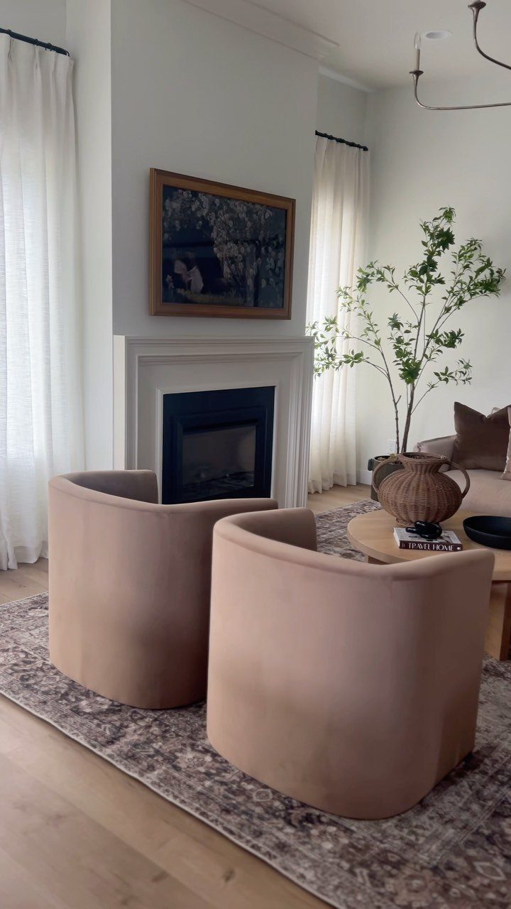 Two beige curved armchairs facing a white fireplace with a painting above it.