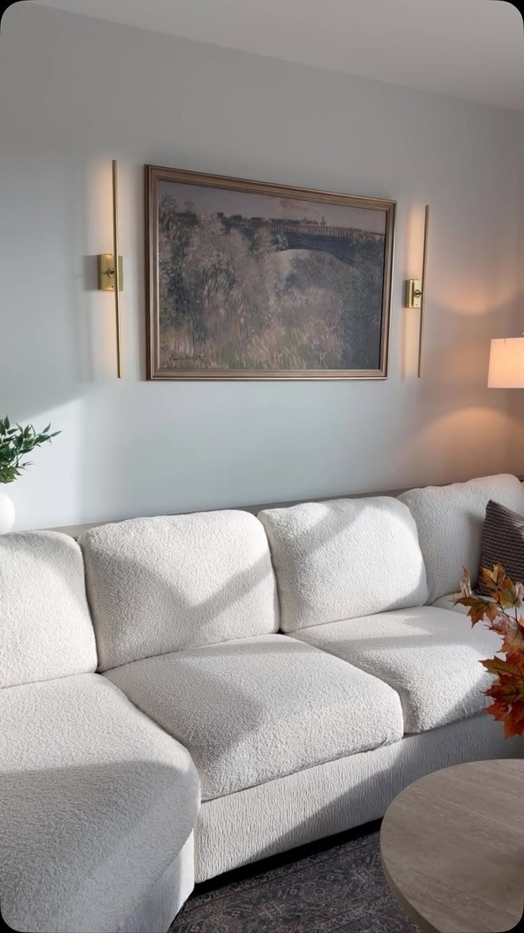 White textured sofa against a grey wall with landscape art and gold sconces.