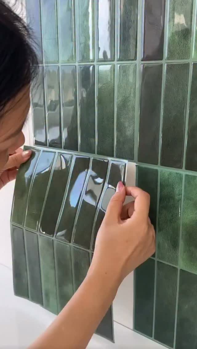 Hands applying green glossy peel-and-stick tiles to a white wall surface.