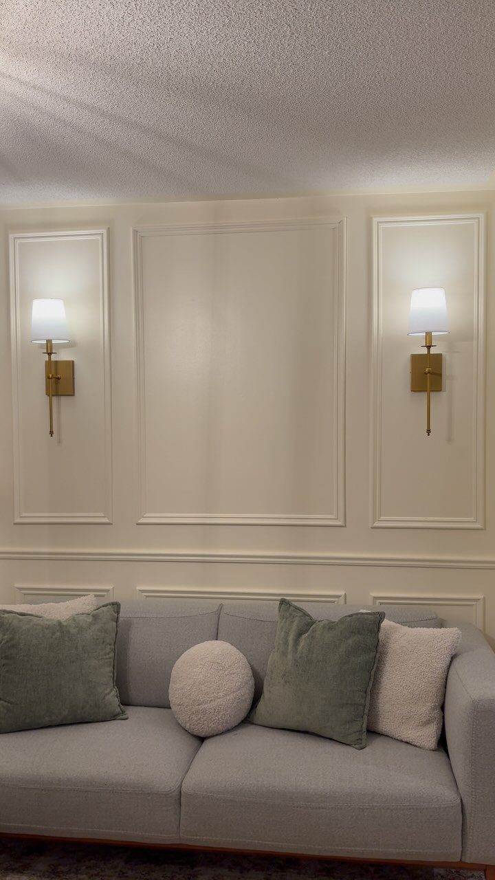 Grey sofa with pillows against a cream wall featuring gold sconces and molding.