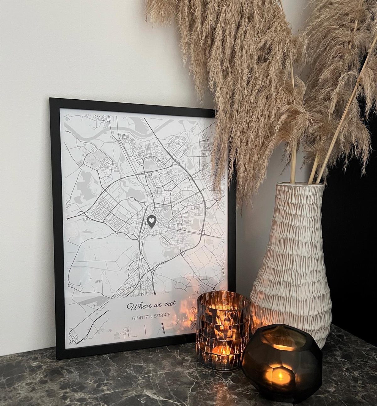Framed city map with heart marker leaning on marble surface with pampas grass.